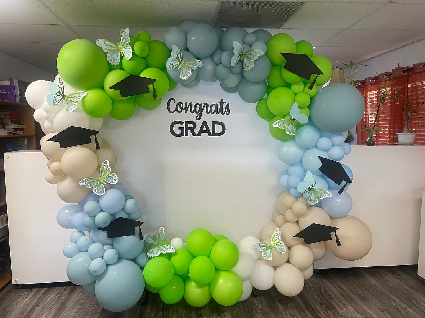 Organic balloon arch in sky blue, lime green, cream, and sand with paper butterflies and graduation-cap accents framing a 'Congrats Grad' wall sign